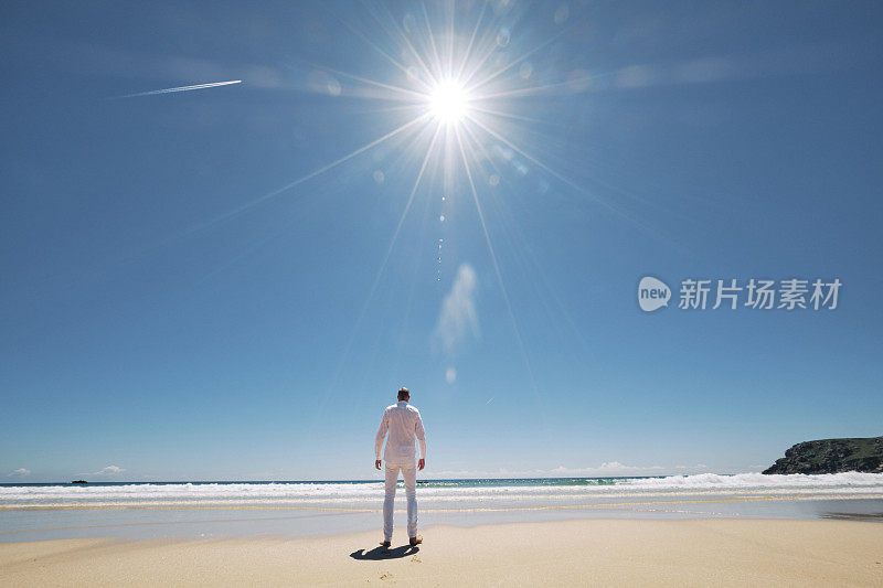 一个身穿白色衣服的男人，独自站在康沃尔的Pedn Vounder海滩的海岸线上，这是一个炎热阳光明媚的六月。
